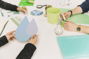 Personas trabajan en una mesa con símbolos de agua, herramientas de oficina y elementos ecológicos que representan sostenibilidad, energía y conciencia ambiental.