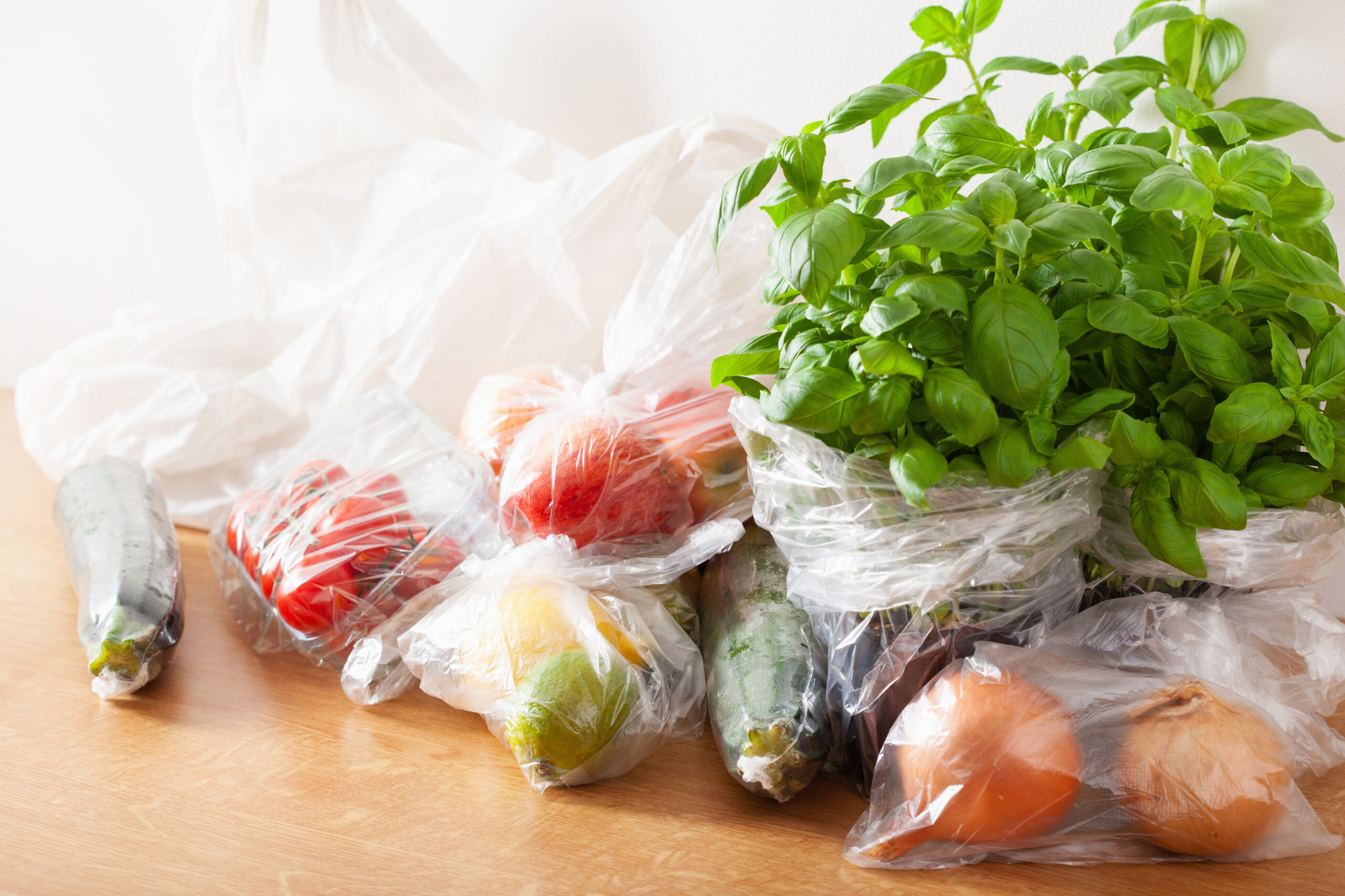 Frutas, verduras y hierbas frescas envueltas en bolsas plásticas de un solo uso sobre una mesa de madera.