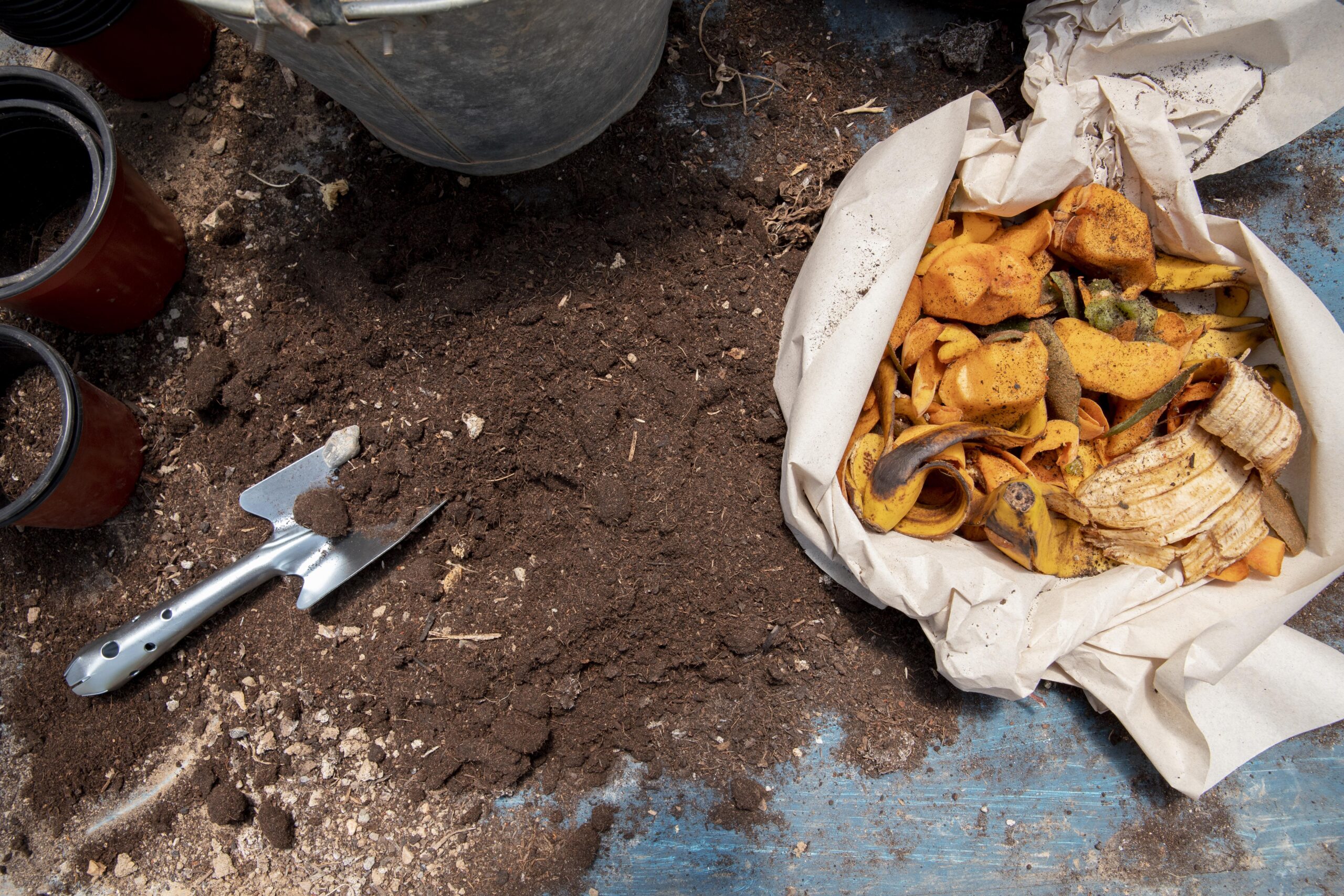 Residuos orgánicos como cáscaras de frutas sobre papel reciclado junto a tierra húmeda y herramientas de jardinería para compostaje.