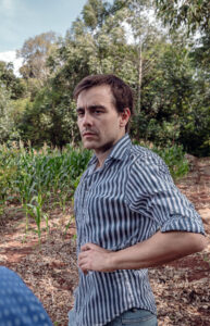 Hombre de camisa a rayas en campo de maíz, rodeado de árboles y suelo rojizo.