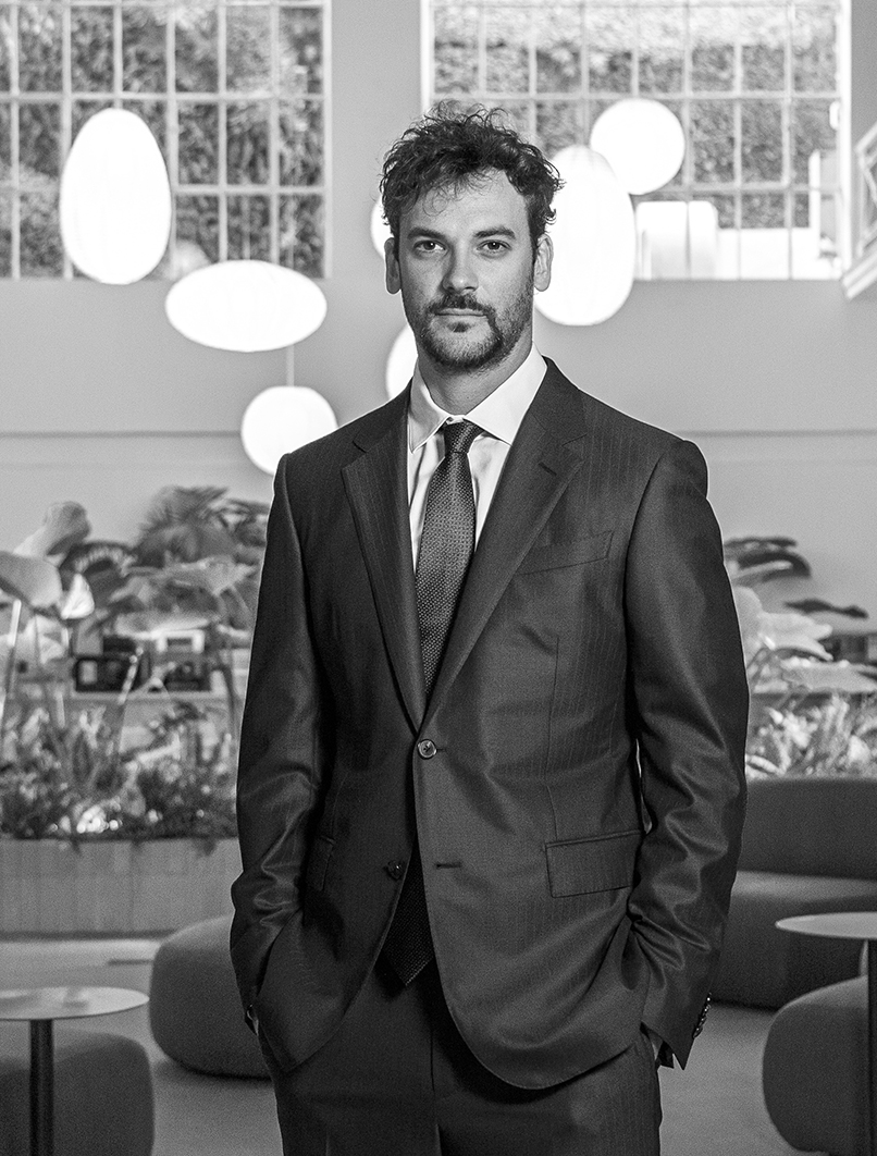 Hombre de traje oscuro posando con expresión seria en un ambiente interior moderno con plantas y luces circulares decorativas.