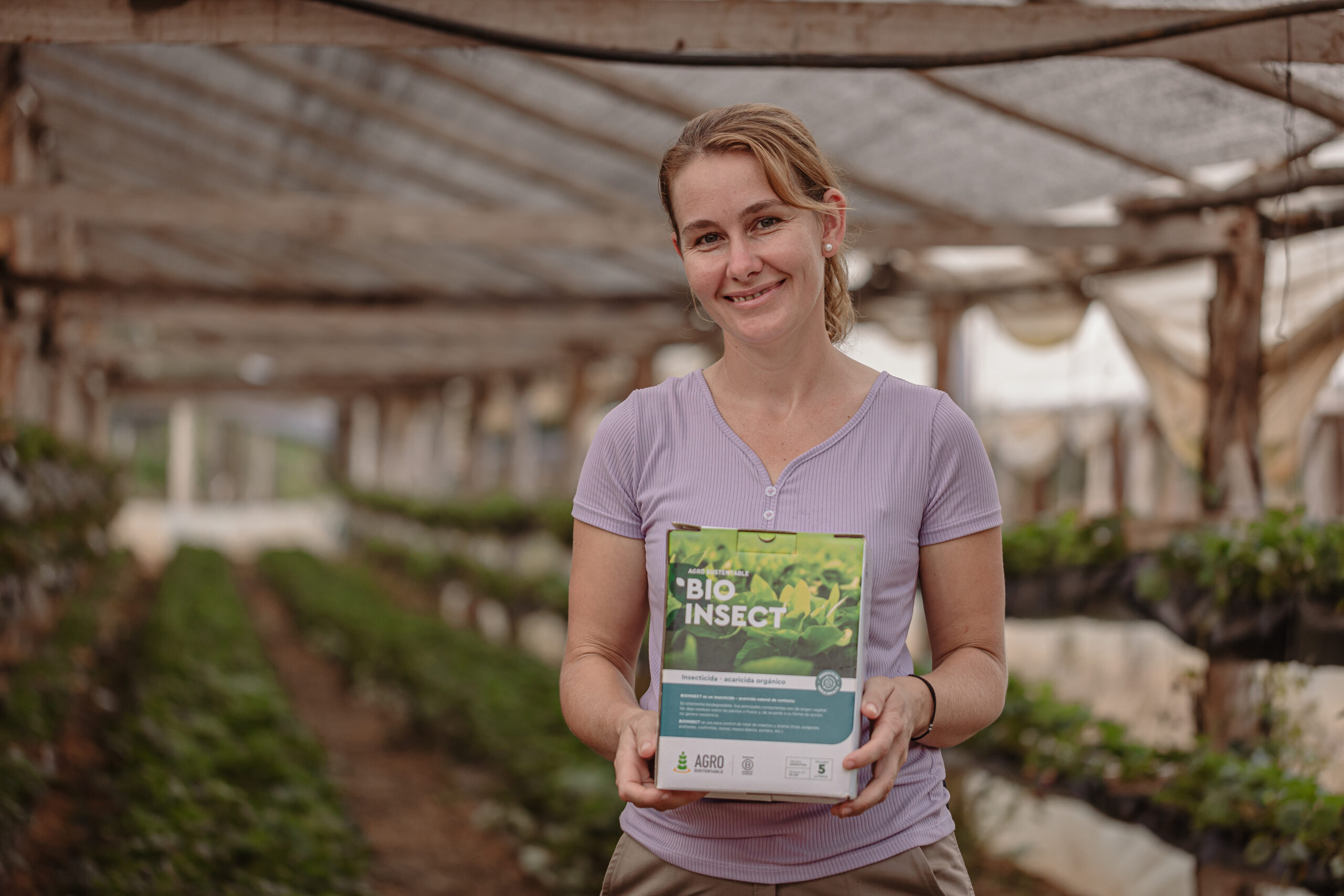 Productora rural sonriente sostiene envase de BioInsect en un invernadero, símbolo de producción sostenible y libre de agroquímicos.
