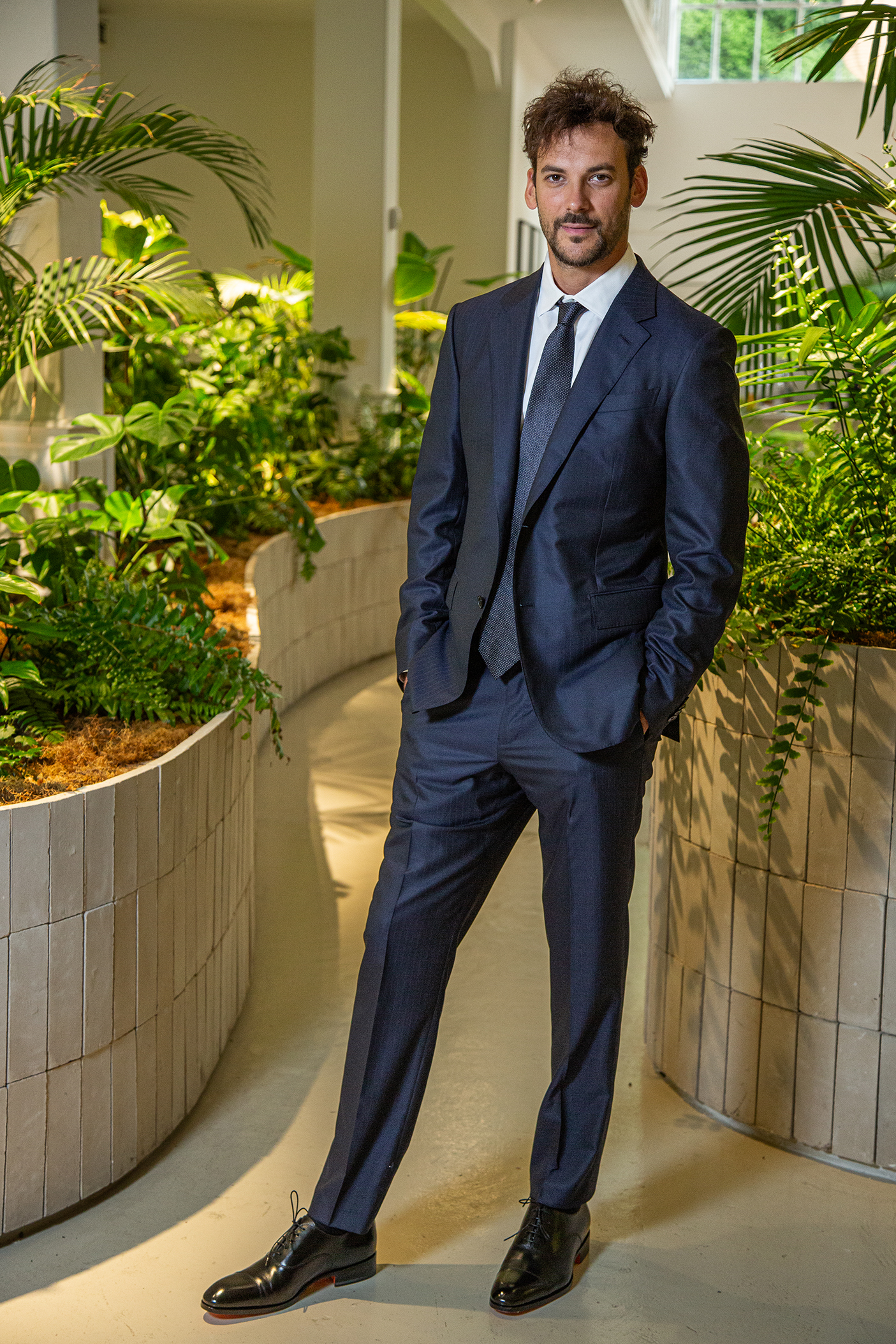 Joaquín Basanta, con traje formal, posa en un entorno natural interior rodeado de plantas tropicales.